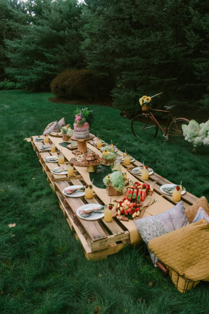 A pallet-wood table on the grass with a small cake, fruit and drinks, a bicycle behind it